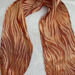 May include: A flowing scarf with a zebra stripe pattern in shades of orange and brown. The scarf is made of a lightweight material and is draped over a white surface.