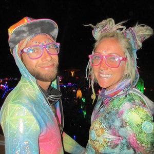 May include: Two people wearing colorful, iridescent clothing and pink-rimmed glasses with rhinestones. The woman has a colorful braided hairstyle and the man is wearing a black and rainbow hooded jacket.