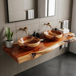 以下が含まれることがあります： 2つの木製の洗面器を備えたバスルームの洗面化粧台。洗面器は、ライブエッジの木製カウンタートップに設置されています。カウンタートップは、豊かな赤茶色です。金色の蛇口と備品が木の色合いを引き立てています。鏡と鉢植えの植物も表示されています。