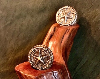 May include: Two gold-toned compass-shaped rings with a star in the center, displayed on a wooden stand.