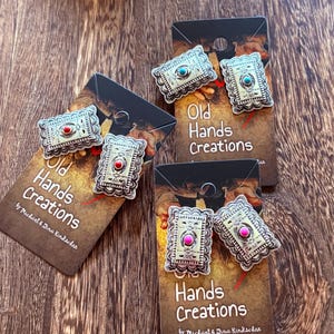 May include: Silver-toned rectangular earrings with a decorative border and a central stone in red, turquoise, or pink. The earrings are displayed on cards that read "Old Hands Creations" and "Hands Creations" by Michael & Dina Kindscher.