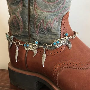 May include: A silver boot charm with turquoise stones and feather accents. The charm is attached to a chain and can be worn on a boot.