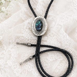 May include: A bolo tie featuring a black braided leather cord and a central oval silver-toned metal slide. The slide contains an iridescent abalone shell inlay. The cord ends are finished with decorative silver-toned metal tips with etched details.
