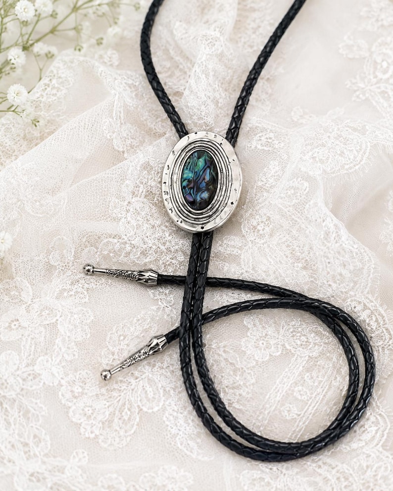 May include: A bolo tie featuring a black braided leather cord and a central oval silver-toned metal slide. The slide contains an iridescent abalone shell inlay. The cord ends are finished with decorative silver-toned metal tips with etched details.