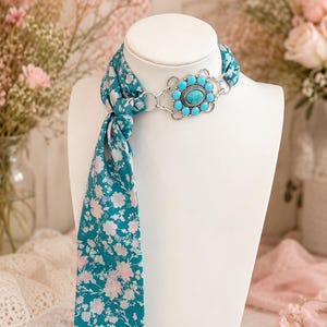 Puede incluir: Un pañuelo de color verde azulado con un estampado floral en blanco y rosa claro atado alrededor de un busto de exhibición blanco. El pañuelo se asegura con un conector de metal plateado con una piedra turquesa ovalada central y cabujones más pequeños.