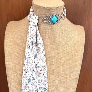 May include: A white floral scarf with pink and blue flowers is draped on a mannequin. The scarf is paired with a silver-toned necklace featuring a turquoise-colored square stone. The necklace is connected to the scarf with silver rings.