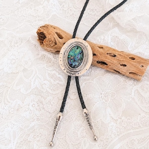 May include: A bolo tie featuring a central oval-shaped abalone shell inlay set within a textured silver-toned metal frame. The cord is made of black braided leather and terminates in two silver-toned metal tips with engraved decorative patterns.