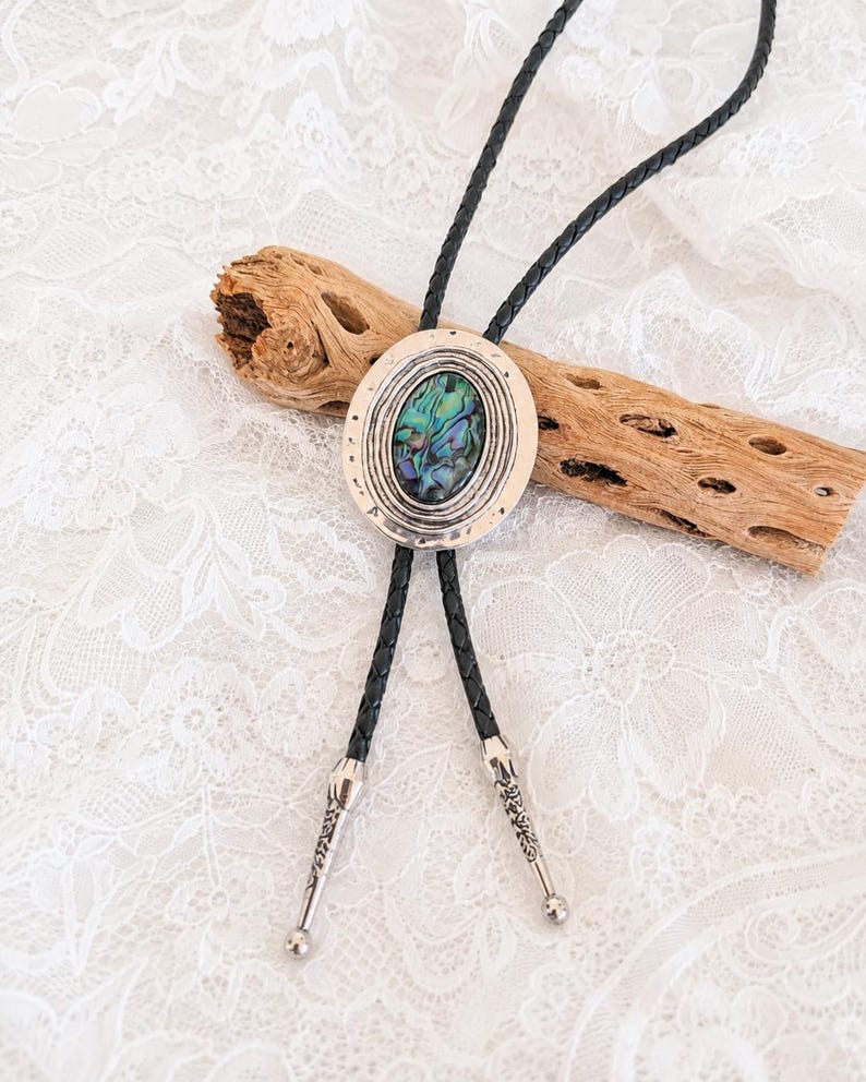 May include: A bolo tie featuring a central oval-shaped abalone shell inlay set within a textured silver-toned metal frame. The cord is made of black braided leather and terminates in two silver-toned metal tips with engraved decorative patterns.