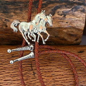 May include: A silver bolo tie featuring two running horses. The horses are intricately detailed and attached to a brown braided leather cord. The tie has decorative silver tips with small ball ends.