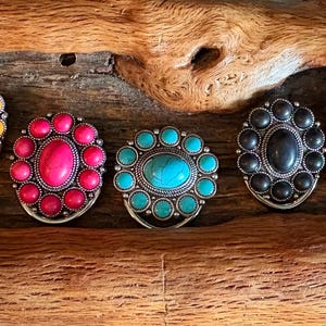 Puede incluir: Seis anillos de metal decorativos con piedras de colores. Cada anillo tiene una piedra ovalada central rodeada de piedras redondas más pequeñas. Las piedras son amarillas, rosas, turquesa, negras y rojas.