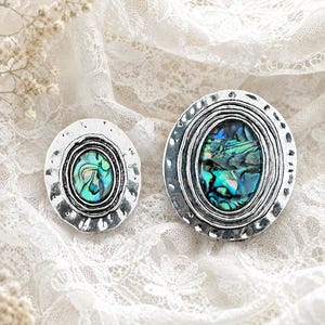 May include: Two oval-shaped brooches featuring iridescent abalone shell inlays set within hammered silver-toned metal frames. Each piece displays a central oval abalone stone surrounded by concentric ridged detailing and a textured outer edge.
