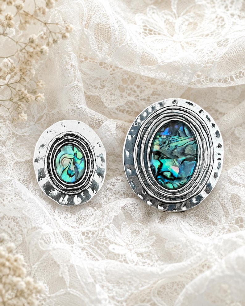 May include: Two oval-shaped brooches featuring iridescent abalone shell inlays set within hammered silver-toned metal frames. Each piece displays a central oval abalone stone surrounded by concentric ridged detailing and a textured outer edge.
