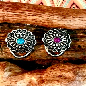 May include: Two silver-toned metal conchos with decorative floral designs. One concho has a turquoise stone in the centre, and the other has a purple stone.