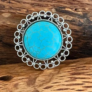 May include: A silver ring featuring a large, round turquoise stone with a cracked pattern. The ring has an ornate, openwork design with small circular details surrounding the central stone. The ring is set against a wooden background.