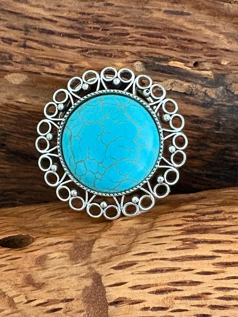 May include: A silver ring featuring a large, round turquoise stone with a cracked pattern. The ring has an ornate, openwork design with small circular details surrounding the central stone. The ring is set against a wooden background.