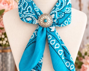 May include: A bright turquoise bandana featuring a white paisley and floral pattern, secured at the center with a round silver-toned metal slide. The slide has a scalloped edge and a small, circular turquoise stone set in the middle.