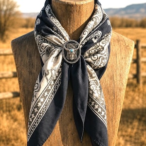 May include: A dark navy blue and cream patterned bandana secured with a silver-toned oval slide featuring a steer skull motif and a small central turquoise stone. The fabric displays a repeating intricate scrollwork design with a geometric border.