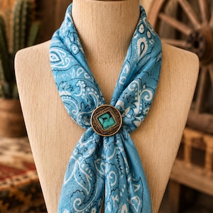 May include: A light blue scarf featuring a white and black paisley pattern, secured with a circular bronze-toned slide fastener that contains a square, marbled turquoise-coloured stone inlay set within a decorative, textured metal frame.