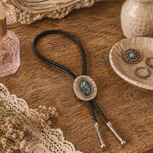 May include: A bolo tie featuring a braided black leather cord and a central oval pendant with an iridescent abalone shell inlay set in a textured silver-tone metal frame. The cord ends are finished with matching engraved silver-tone metal tips.