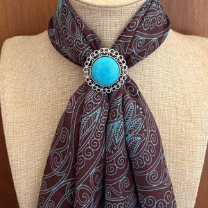 May include: A brown scarf with a teal and blue paisley design is fastened with a silver-coloured bolo tie. The bolo tie features a large, round, turquoise-coloured stone in the centre, surrounded by a decorative silver border. The scarf is draped over a mannequin.