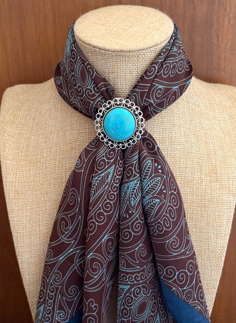 May include: A brown scarf with a teal and blue paisley design is fastened with a silver-coloured bolo tie. The bolo tie features a large, round, turquoise-coloured stone in the centre, surrounded by a decorative silver border. The scarf is draped over a mannequin.