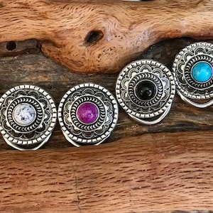 May include: Six silver rings with different colored stones in the center. The stones are red, white, purple, black, blue, and turquoise. The rings are on a wooden surface.