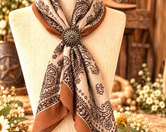 May include: A beige and brown paisley print scarf secured with a round, metallic, sunburst-patterned slide. The scarf features a dark brown border and is draped over a mannequin, showcasing the intricate dark brown paisley motifs on a light beige fabric.