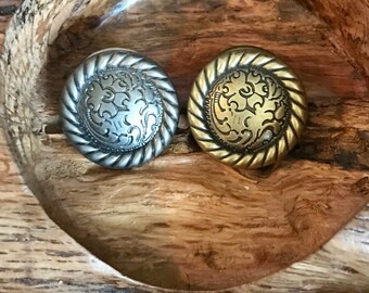 May include: Two decorative metal buttons with a rope-like border and a floral design. The button on the left is silver and the button on the right is gold.