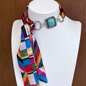 May include: A colorful neck scarf with a square turquoise stone pendant. The scarf features a vibrant patchwork design in various colors, including red, blue, green, and yellow. The pendant is set in a silver-toned frame.