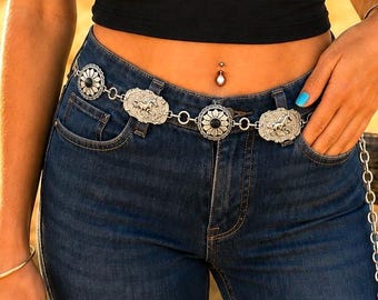 May include: A silver-toned chain belt featuring alternating circular and rectangular metal plates. The circular plates contain a central black stone surrounded by a floral pattern, while the rectangular plates feature an embossed horse motif.