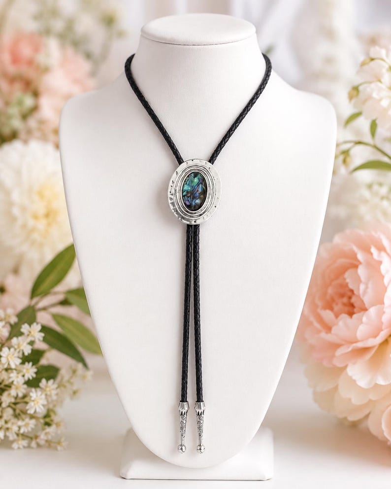 May include: A bolo tie featuring a central oval pendant with a shimmering iridescent abalone shell inlay set in a textured silver-tone frame. The pendant is attached to a black braided cord finished with two matching silver-tone metal tips.