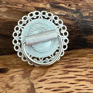 May include: A silver drawer pull with a circular design. It has an openwork border around a central, light-coloured image. A small, cylindrical handle extends from the back, with a curved, silver handle attached.