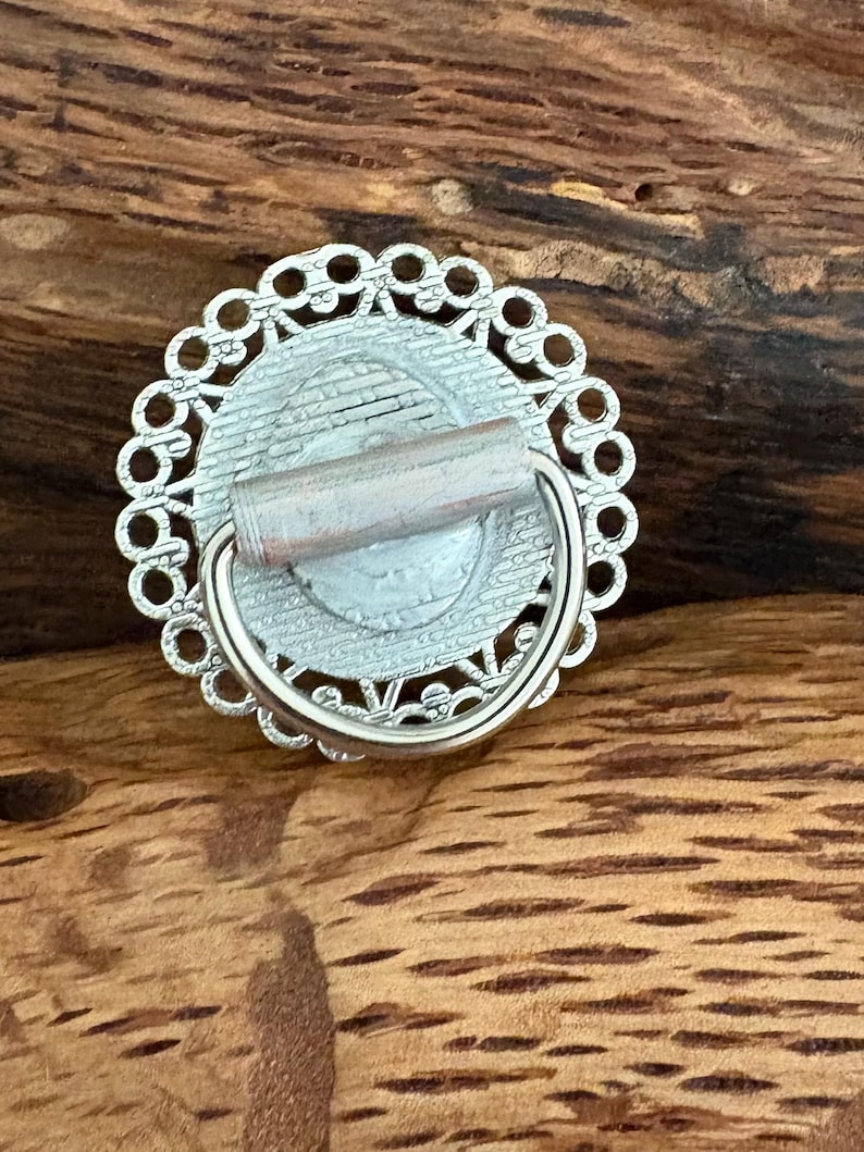 May include: A silver drawer pull with a circular design. It has an openwork border around a central, light-coloured image. A small, cylindrical handle extends from the back, with a curved, silver handle attached.