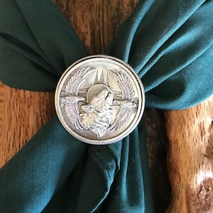 May include: A silver-toned metal neckerchief slide with a wolf howling at the moon design. The slide is on a dark green neckerchief.
