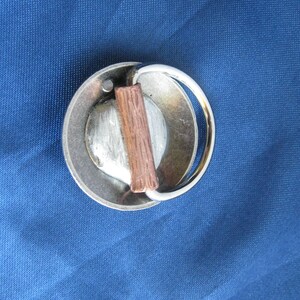 May include: A silver metal belt buckle with a wooden insert. The buckle has a rounded shape and a ring for attaching a belt.
