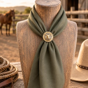 May include: An olive green scarf secured with a circular brass slide featuring a central bull skull motif. The slide has a textured, decorative border surrounding the bull head, holding the fabric in a neat, draped knot at the center of the scarf.