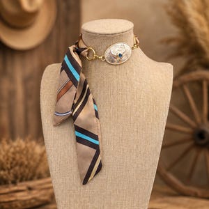 May include: A choker necklace featuring a tan, brown, and bright blue striped silk scarf tied to a gold-toned metal ring. The centerpiece is an oval-shaped silver-toned metal pendant with engraved scrollwork and a small, round blue stone.