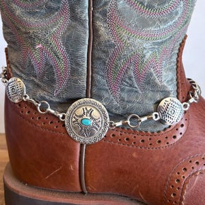 May include: A silver chain with round turquoise and silver charms attached to a brown leather boot.