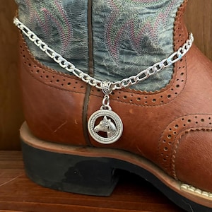 May include: A silver boot chain with a horse head pendant is draped over a brown leather cowboy boot. The boot has a decorative blue and green patterned shaft. The chain is made of small, interlocking links.