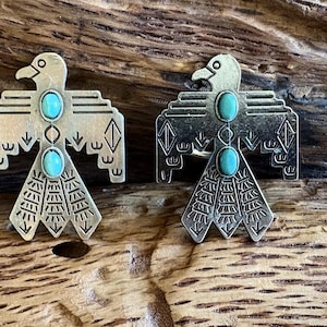 Op de afbeelding: Twee zilverkleurige metalen vogelvormige hangers met turquoise ovale stenen. De hangers hebben gedetailleerde vleugel- en staartontwerpen. De achtergrond is een getextureerd houten oppervlak.