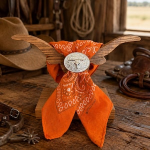 May include: A wooden display stand shaped like longhorns holds an orange bandana with a black paisley pattern. A silver-toned oval concho featuring a steer skull design secures the fabric at the center of the wooden horns on the display base.