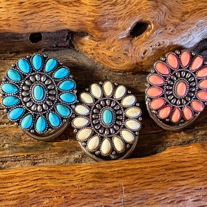 Puede incluir: Tres adornos redondos decorativos de metal y piedra. Los adornos son en un esquema de colores turquesa, blanco y coral. Los adornos tienen un diseño circular con una piedra central rodeada de piedras más pequeñas.