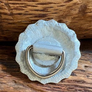 May include: A circular silver-toned metal drawer pull featuring a textured, scalloped edge and a central sunburst pattern. A smooth, semi-circular metal ring handle is attached to the center, allowing for a pivoting motion when pulled.