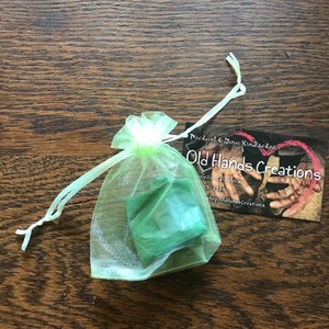 May include: A small, light green organza gift bag with a drawstring closure. Inside the bag is a folded green item. A business card with the text "Old Hands Creations" and contact information is next to the bag.