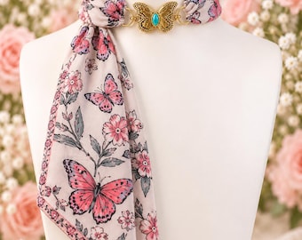 May include: A light pink scarf featuring a floral and butterfly print, secured at the neck with a gold-toned metal butterfly clasp. The clasp is centered and features a small, oval-shaped turquoise stone set in the middle of the butterfly wings.