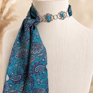 May include: A teal scarf featuring a dark blue and white paisley pattern, secured by a silver-toned metal choker necklace. The necklace consists of circular links and two oval-shaped ornaments, each set with a smooth, bright blue stone.