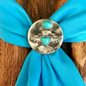 May include: A turquoise blue bandana with a silver bolo tie featuring a turquoise heart and a bird design.
