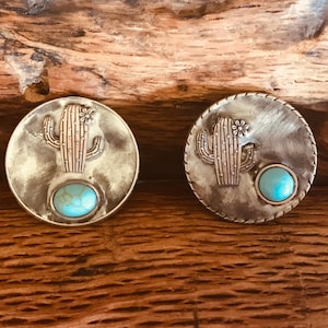 Puede incluir: Dos botones redondos de metal plateado con una piedra turquesa y un diseño de cactus.