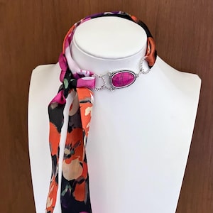 May include: A floral scarf necklace with a silver-toned clasp and a pink stone. The scarf features a colorful floral pattern with shades of pink, orange, and black. The necklace is displayed on a white mannequin.