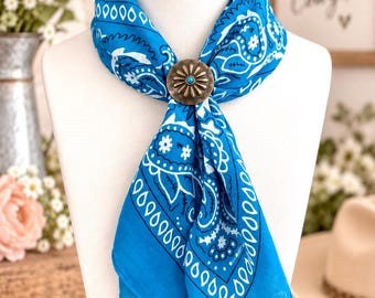 May include: A bright blue bandana featuring a white paisley and floral pattern, secured at the center with a round, antique-finish metal slide. The slide has a sunburst design with a small, central turquoise-colored stone accent.